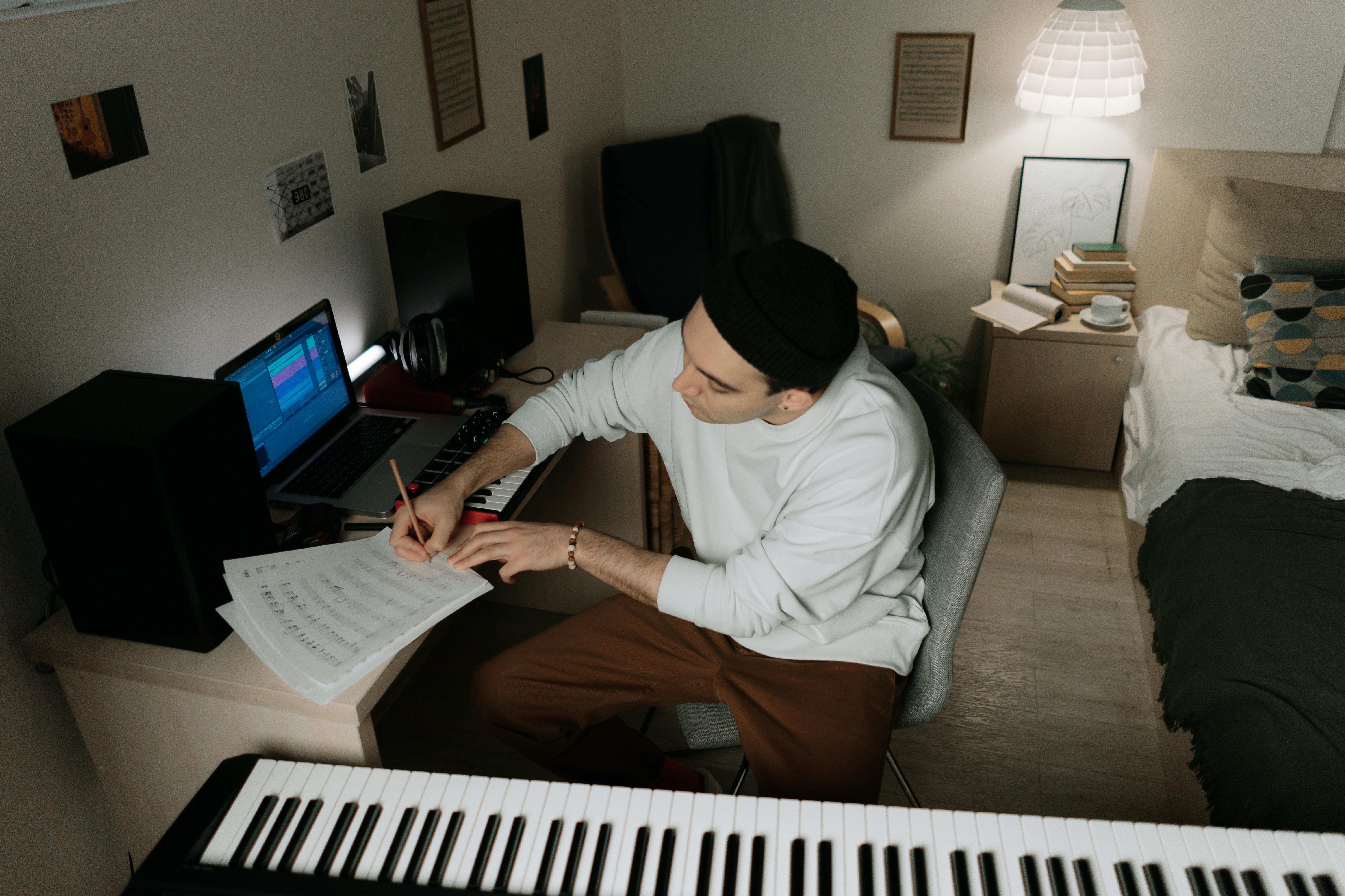 Homme jouant du piano dans sa chambre, écrivant sur une feuille, avec un ordinateur à côté.