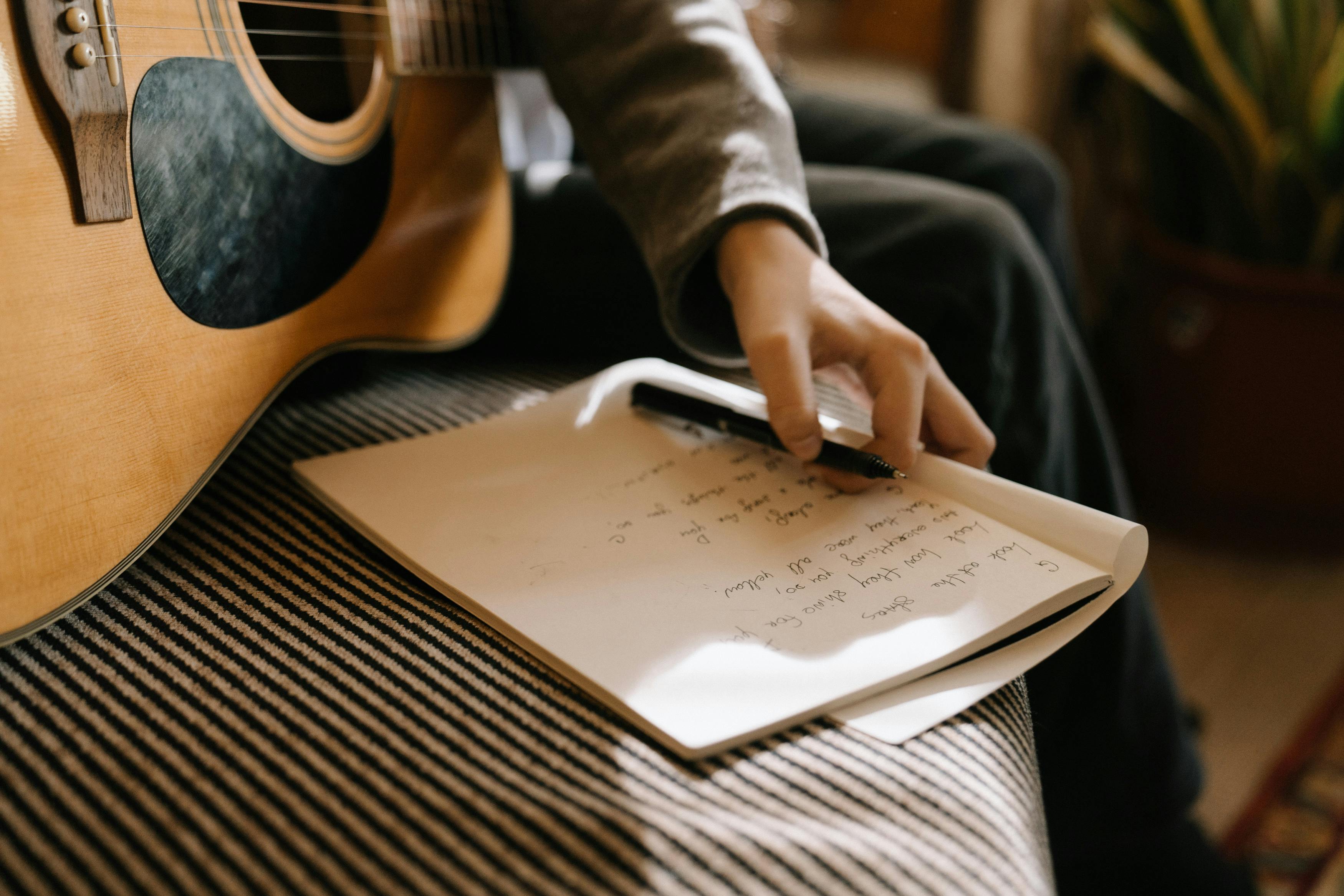 Main tenant une guitare, avec une feuille remplie de notes et un stylo posé à côté.