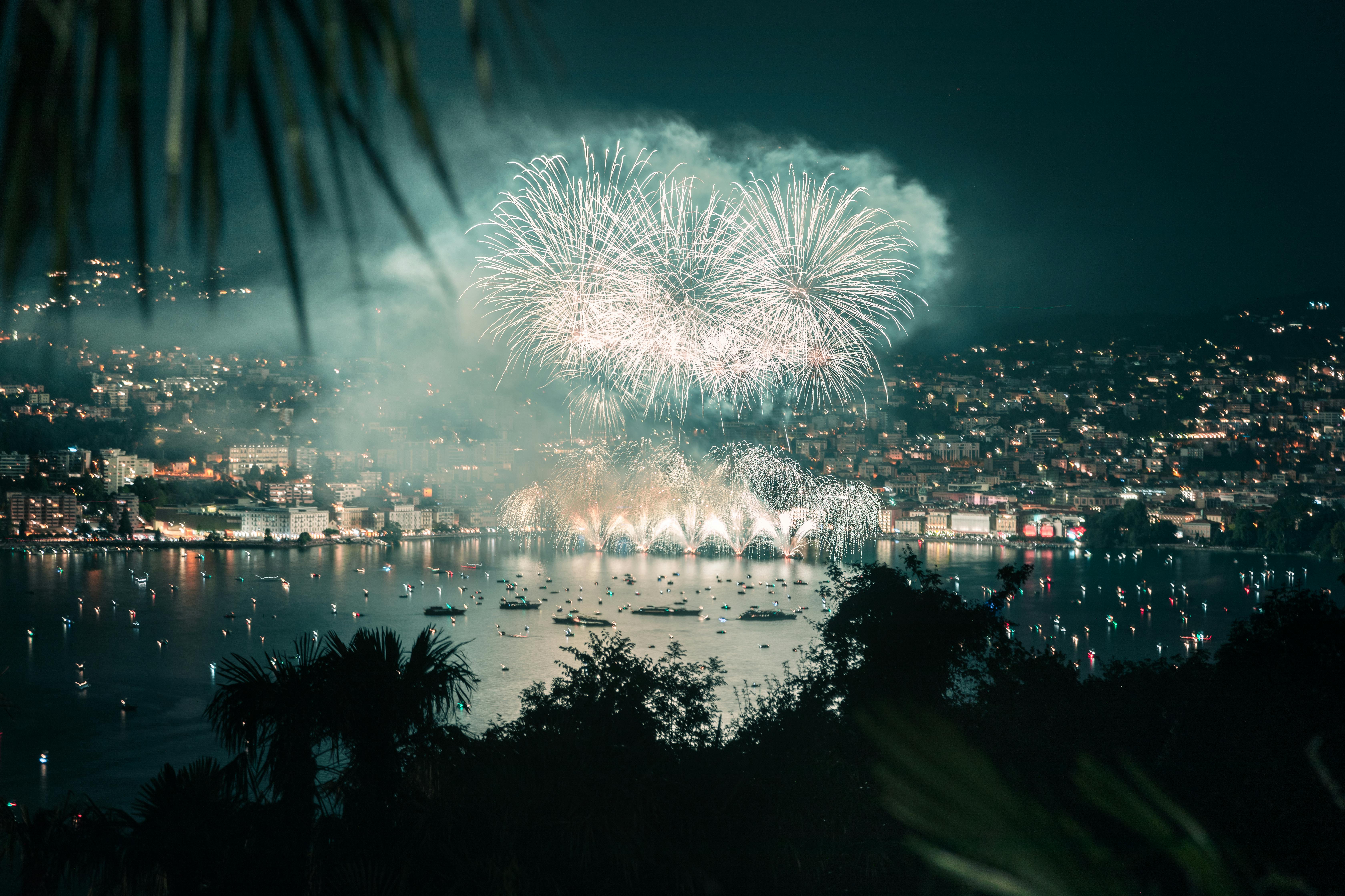 Un feu d’artifice spectaculaire éclate au-dessus de l’eau, illuminant bateaux et bâtiments alentour, dans un décor urbain féerique de nuit