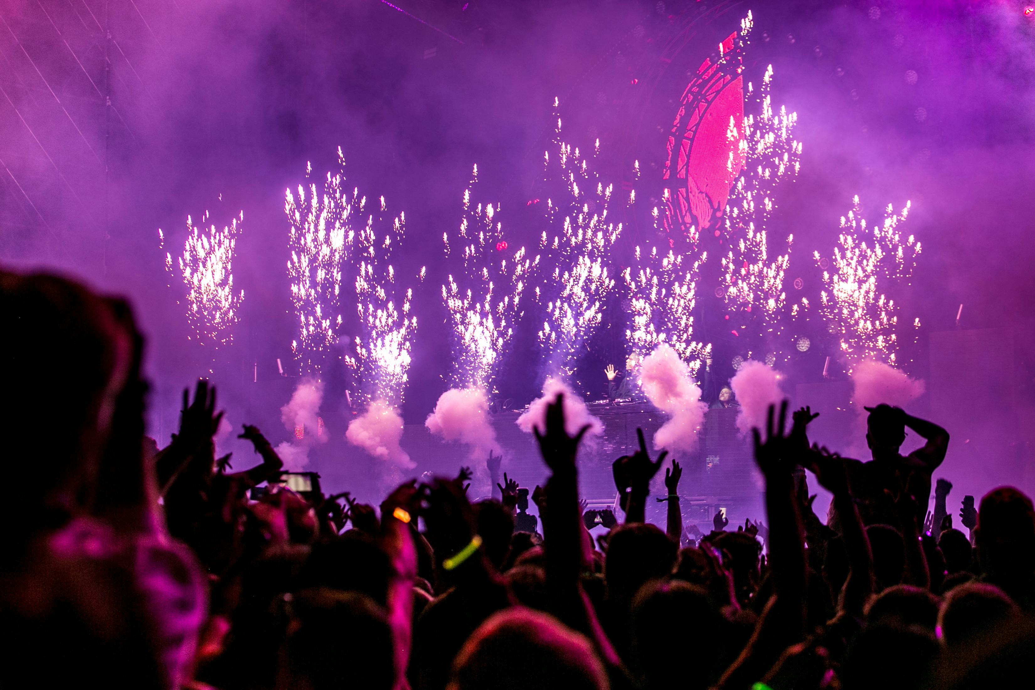 Foule en liesse devant une scène illuminée de feux d’artifice et de fumée violette, les mains levées et les téléphones brandis dans une ambiance festive
