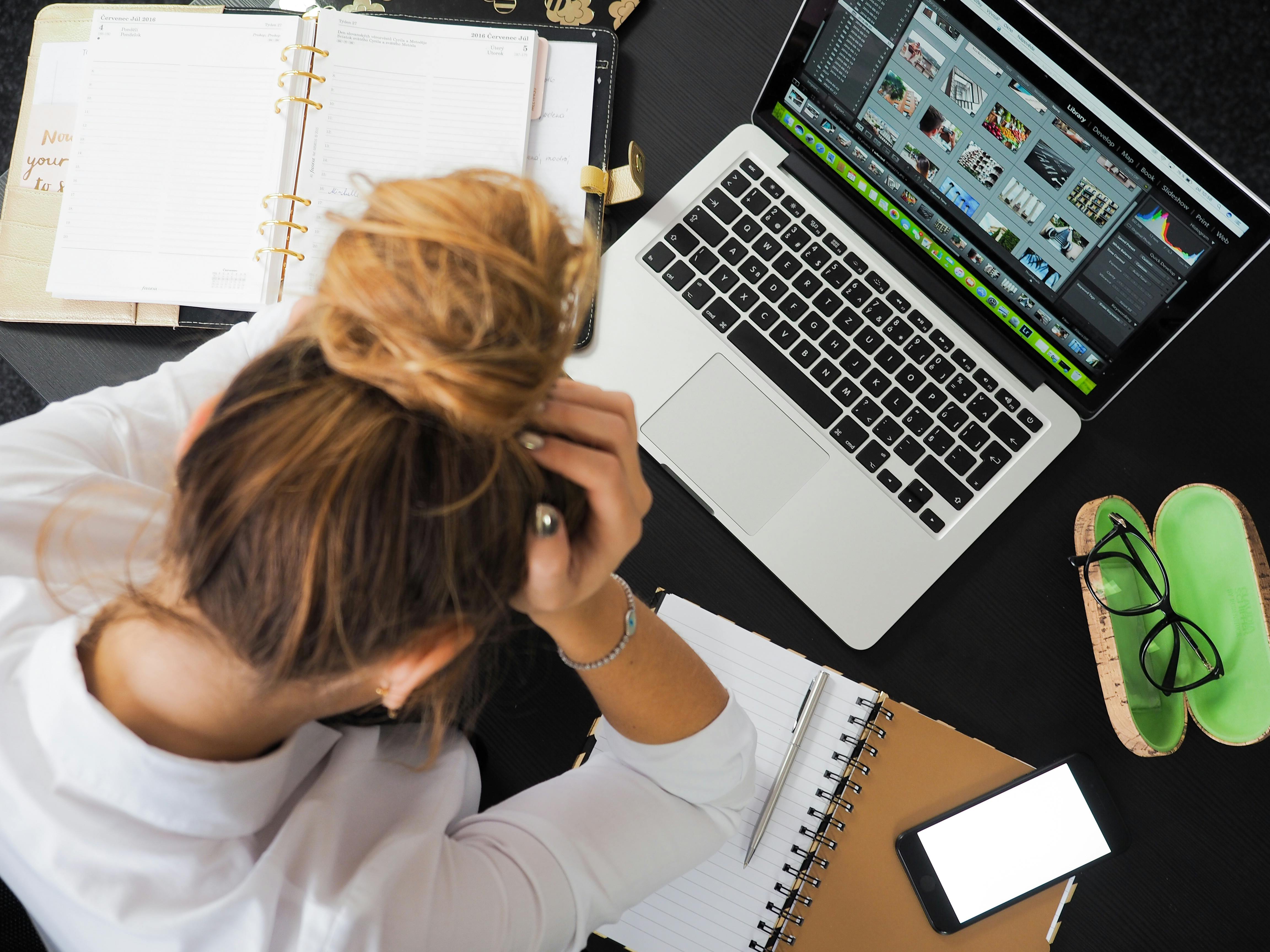 Femme, la tête entre les mains, face à un bureau encombré d’ordinateur, cahiers, stylos et téléphone