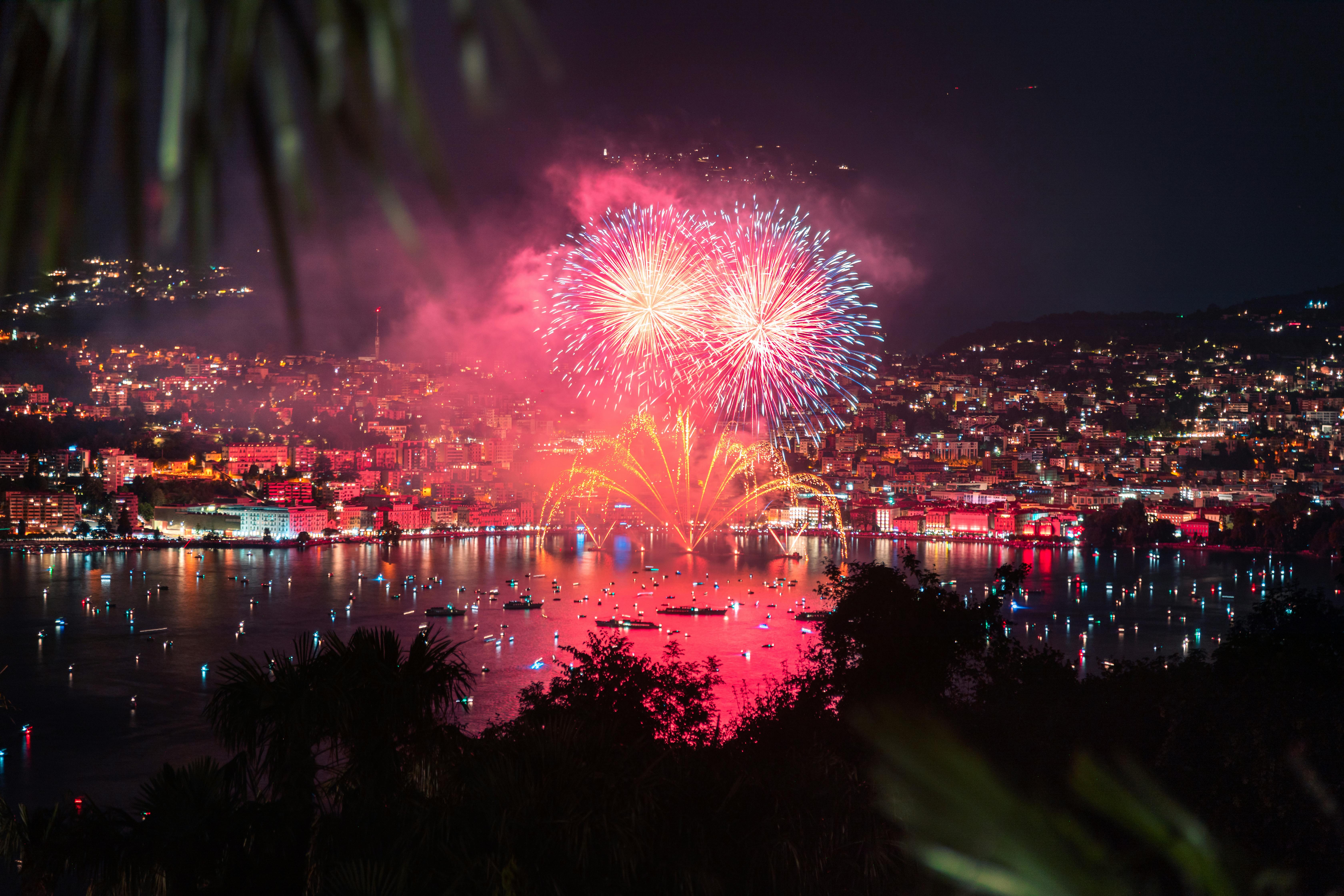 Un vaste plan d'eau calme, entouré de verdure en premier plan de ville au second, avec, reflétant le ciel éclairé par des feux d’artifice 