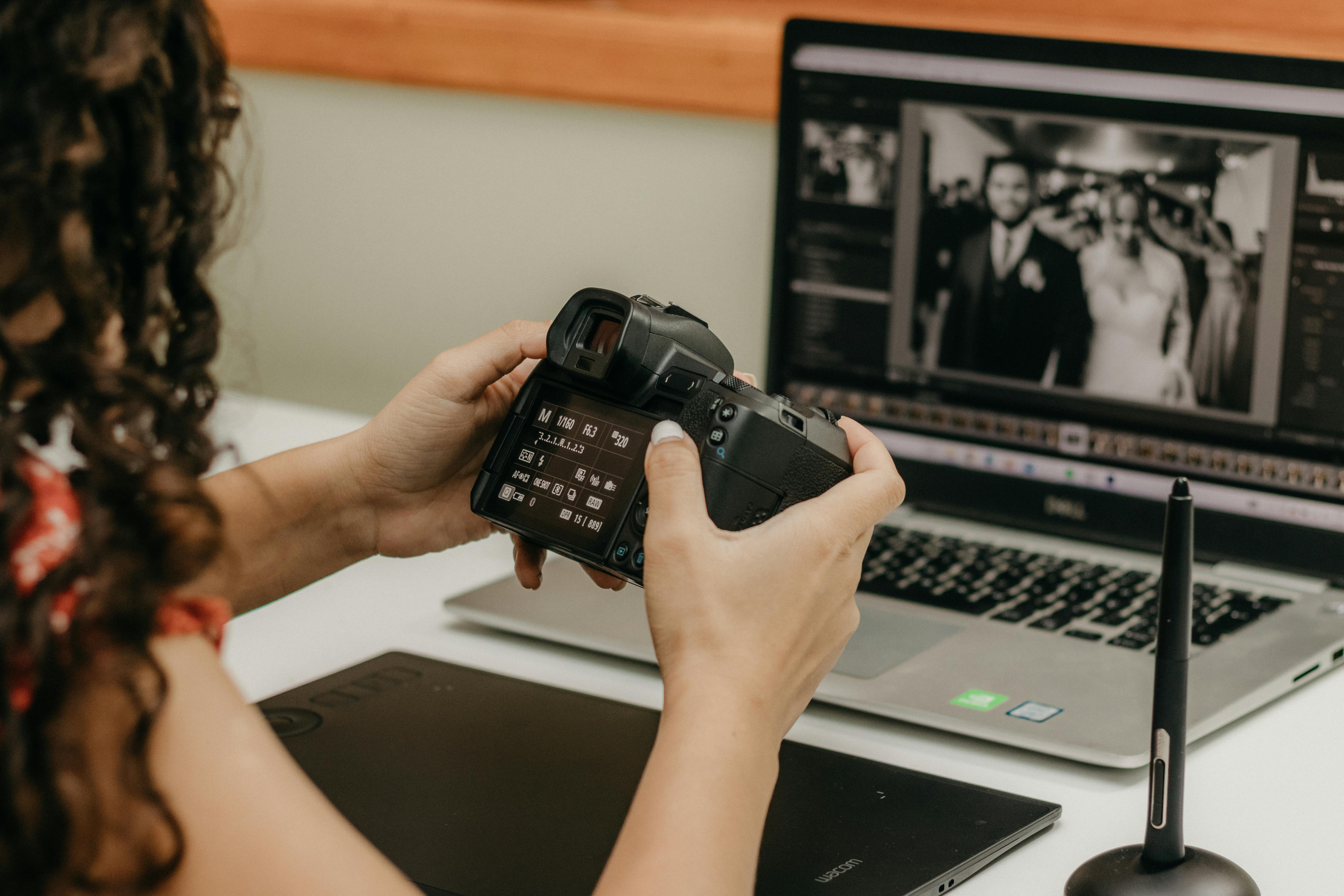 Femme effectuant des réglages photo sur un ordinateur, avec un appareil photo dans les mains