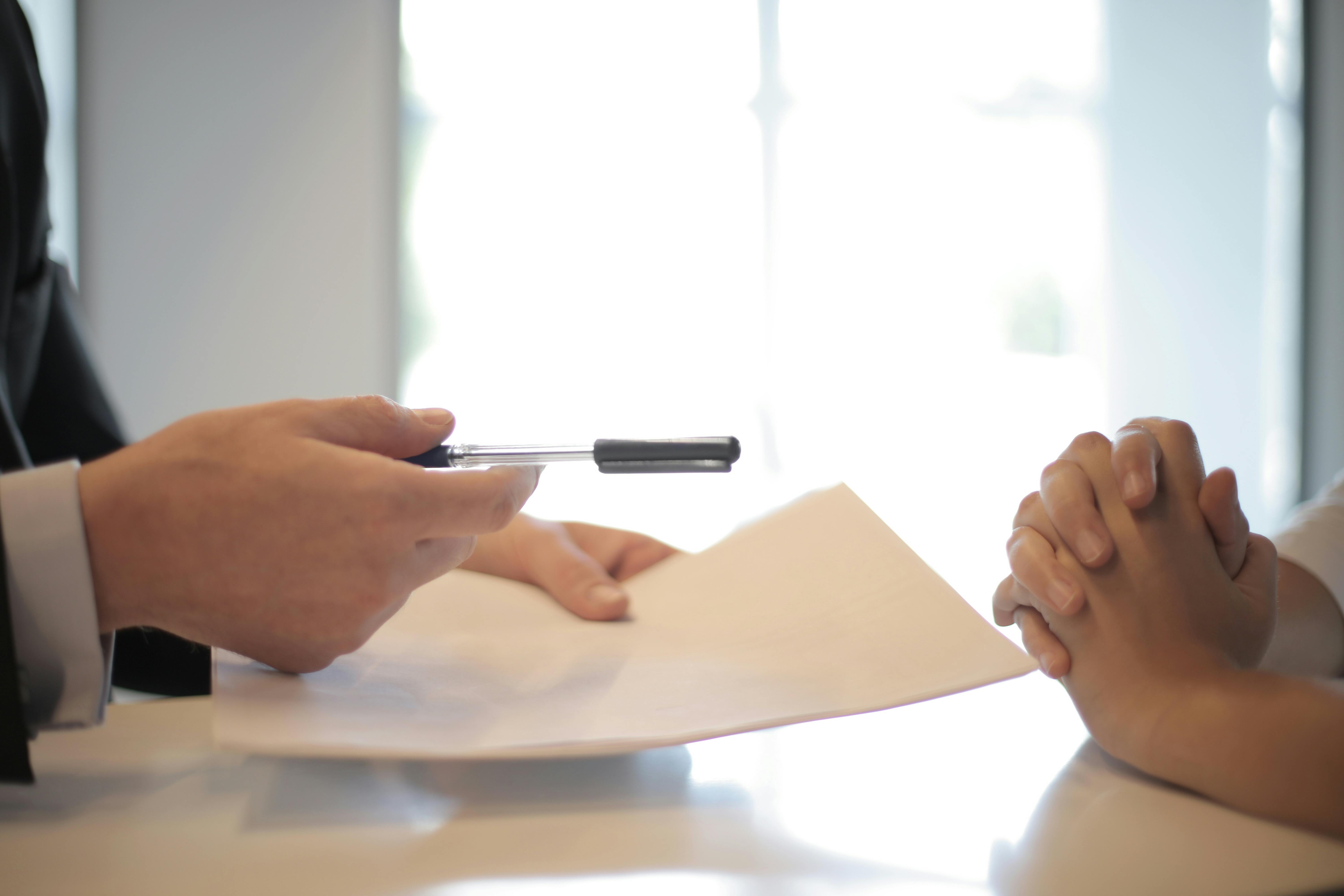 Un homme et une femme assis à une table, l’homme tendant un stylo à la femme pour signer, remplir un papier
