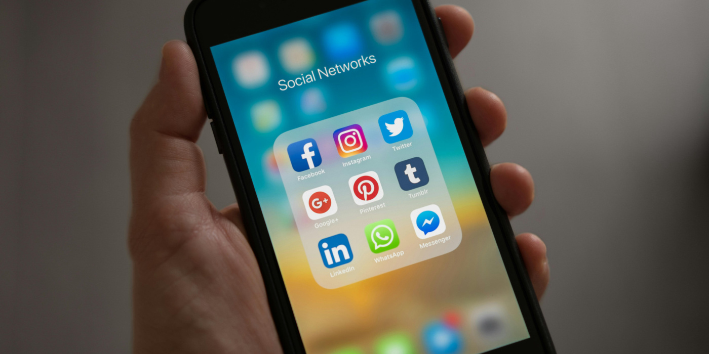 Main tenant un smartphone affichant les icônes des principales applications de réseaux sociaux : Facebook, Instagram, Twitter, LinkedIn, WhatsApp, Messenger, Pinterest et autres